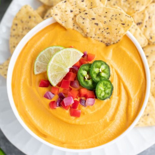 A bowl of yellow melty vegan cheese dip topped with limes, chopped red peppers, red onions, and jalapenos