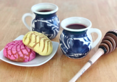 2 cups of champurrado in white mugs with a blue design, sitting on top of a wooden table next to a plate of Mexican pastries and a Mexican whisk called a molinillo