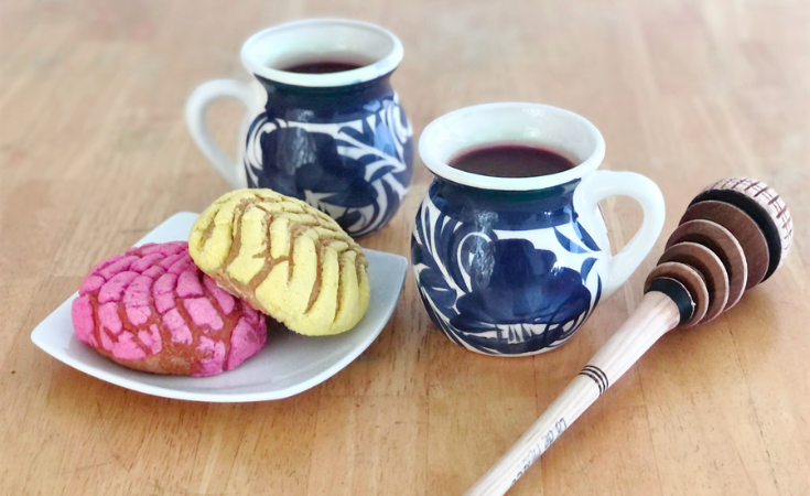 2 cups of champurrado in white mugs with a blue design, sitting on top of a wooden table next to a plate of Mexican pastries and a Mexican whisk called a molinillo