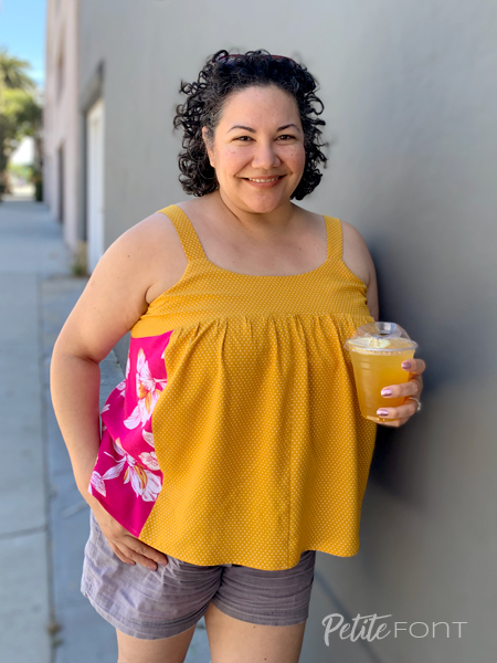 Paulette holding a cold drink leaning up against a grey wall in a pink and gold Marcel tank