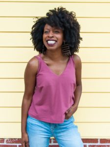 Black woman smiling off camera in a pink Misty camisole over light wash jeans, against a yellow wall